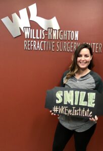 SMILE Shreveport Patient posing for a photo in front of the "Willis Knighton Refractive Surgery Center. They're holding a sign that reads"SMILE #WKEyeInstitute"